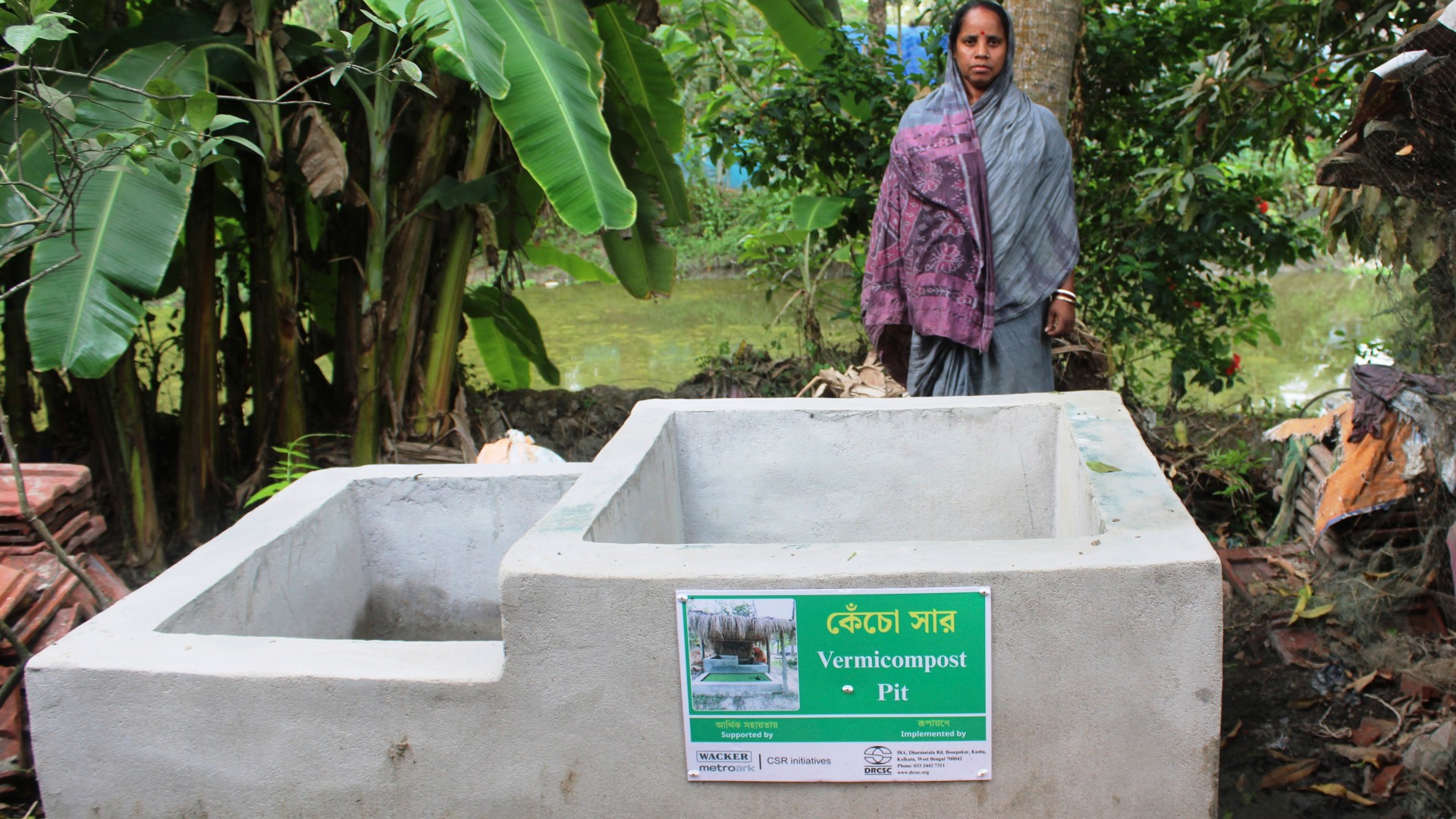 A woman in traditional Indian clothing stands behind a compost pit. A sign on the compost pit indicates that the irrigation system is supported by Wacker Metroark.