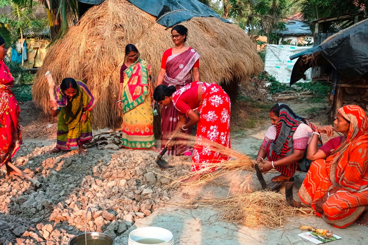 Several smiling women in colorful saris work together on a field, preparing it for cultivation