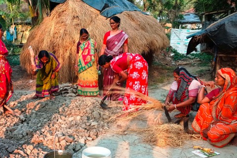 Several smiling women in colorful saris work together on a field, preparing it for cultivation