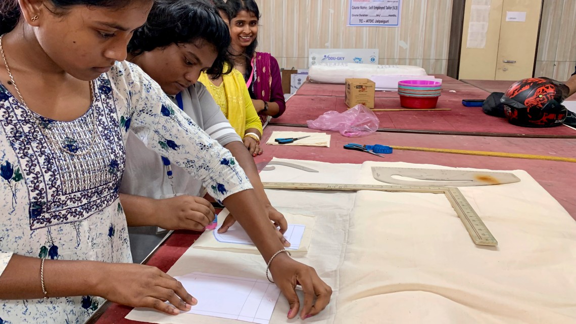 Two women are concentrating on drawing patterns onto a light-colored fabric; two other women are standing nearby, smiling and giving instructions.