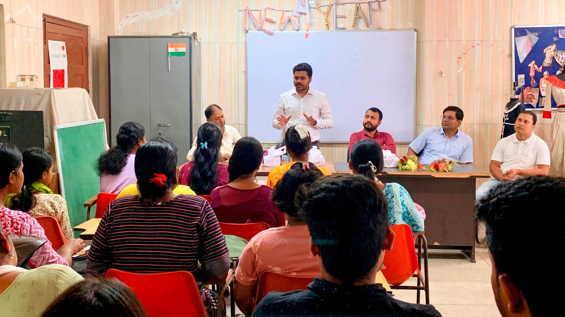 An instructor stands at a table in a classroom and speaks to the trainees sitting in front of him.