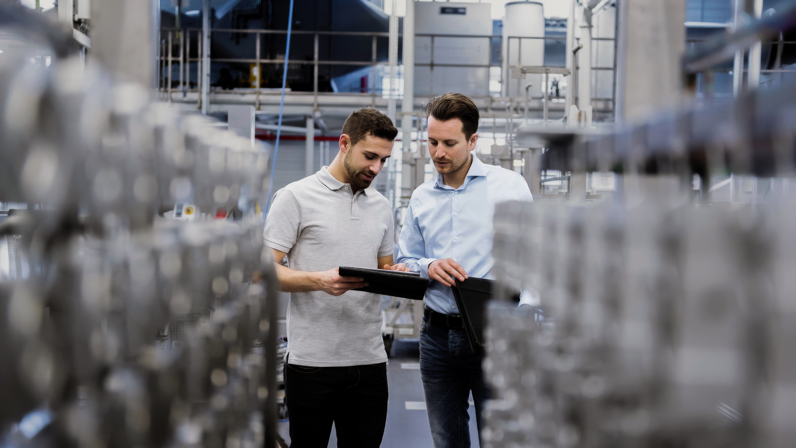 Zwei M&auml;nner in Businesskleidung stehen in einer Produktionshalle und schauen auf ein Tablet.