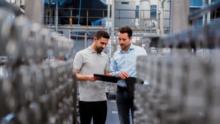 Zwei M&auml;nner in Businesskleidung stehen in einer Produktionshalle und schauen auf ein Tablet.