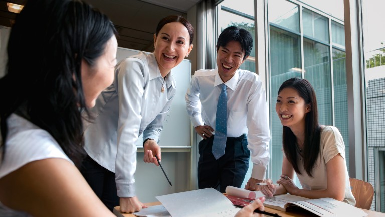 Vier Personen sitzen und stehen in einer Arbeitssituation um einen Schreibtisch herum und lachen.