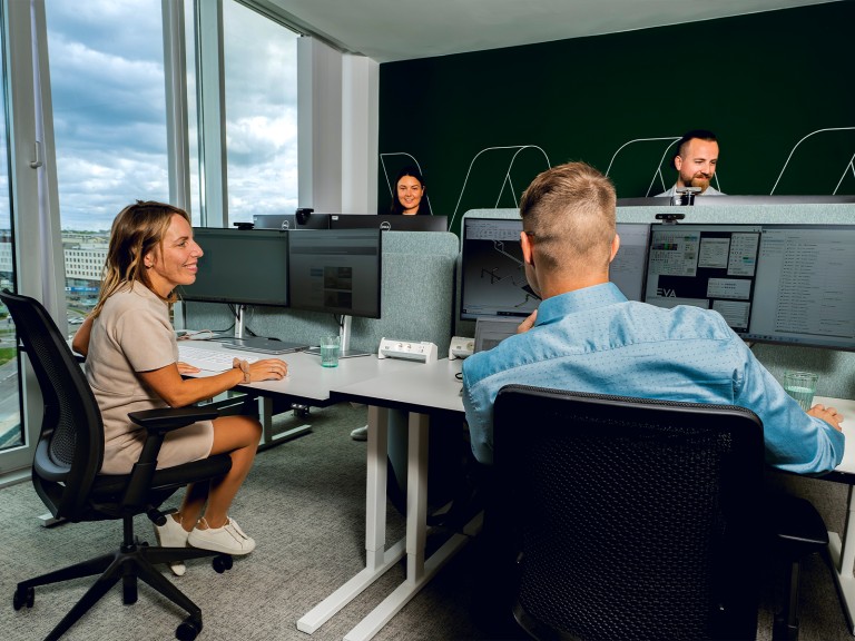 Vier Personen sitzen mit fr&ouml;hlichen Gesichtern in einem B&uuml;ro an ihren Schreibtischen.