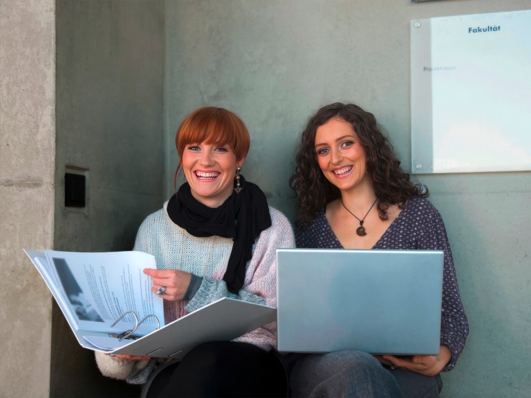 Zwei Studentinnen mit Laptop und Ordner l&auml;cheln in die Kamera.