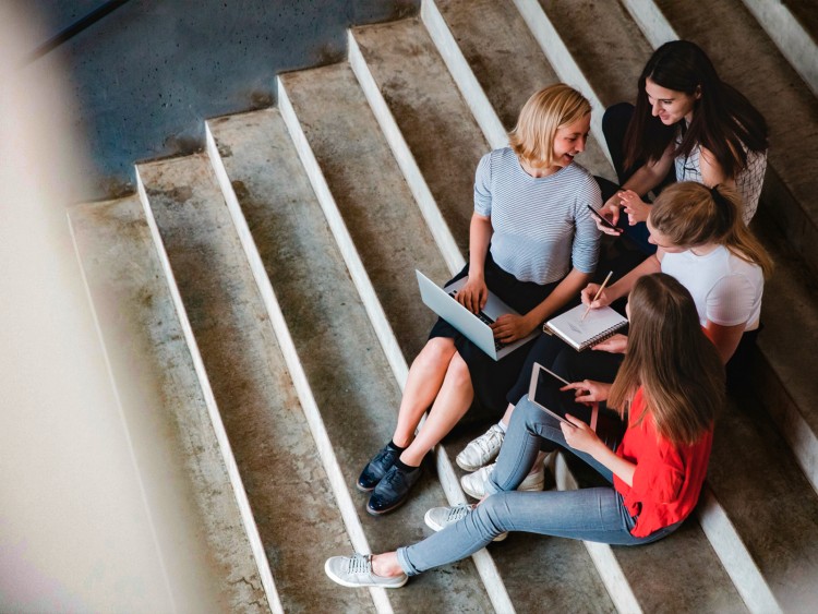 Vier Studentinnen sitzen mit Laptop, Tablet, Handy und Notizheft auf einer Treppe und unterhalten sich.