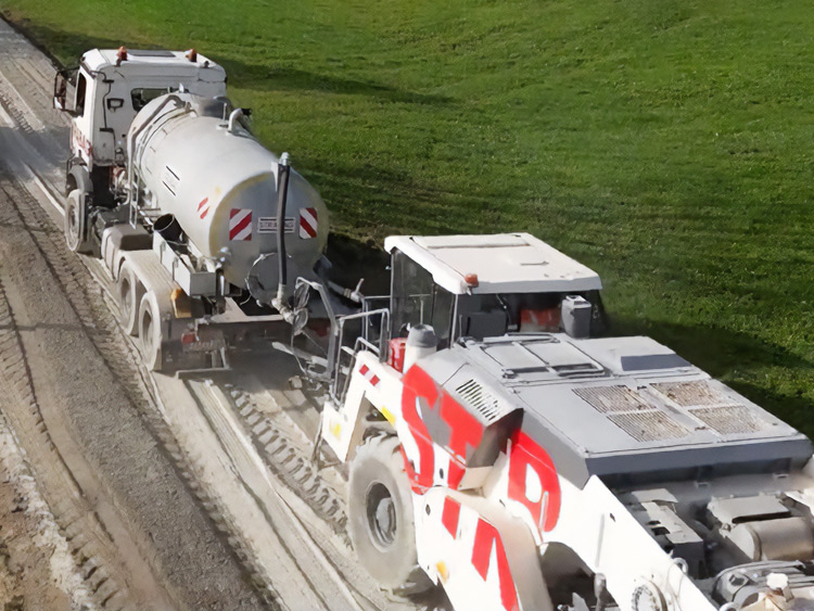 Road construction machinery is mixing a liquid binder with cement into the ground on a country road; the water truck is supplying the recycler with the binder diluted in water.