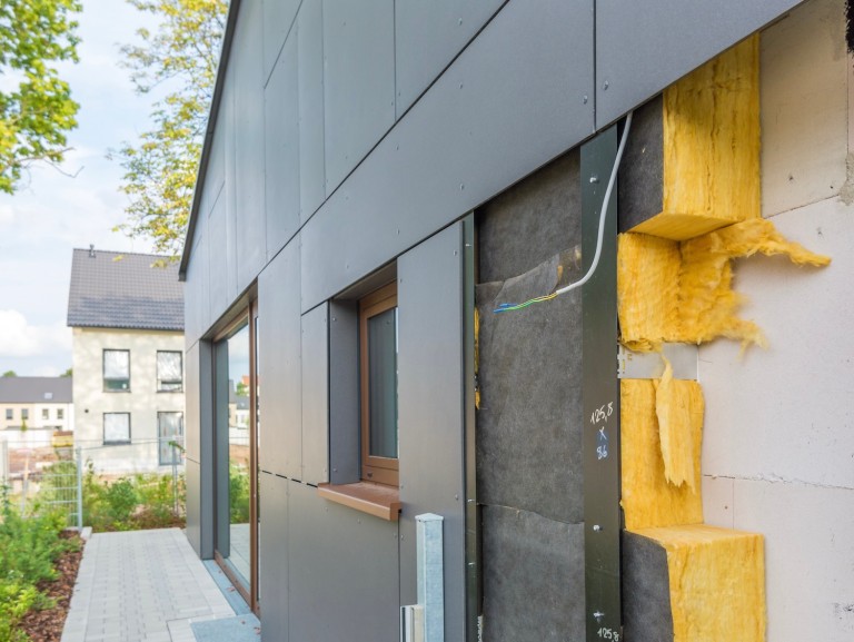 Photo of a modern house facade with visible thermal insulation: the cut-away exterior wall shows yellow wool insulation boards, substructure, and facade cladding.