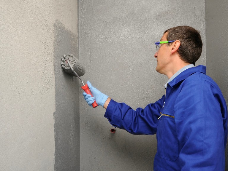 Professional installer applying a polymer-modified waterproofing membrane with a roller in a bathroom as tile underlayment.